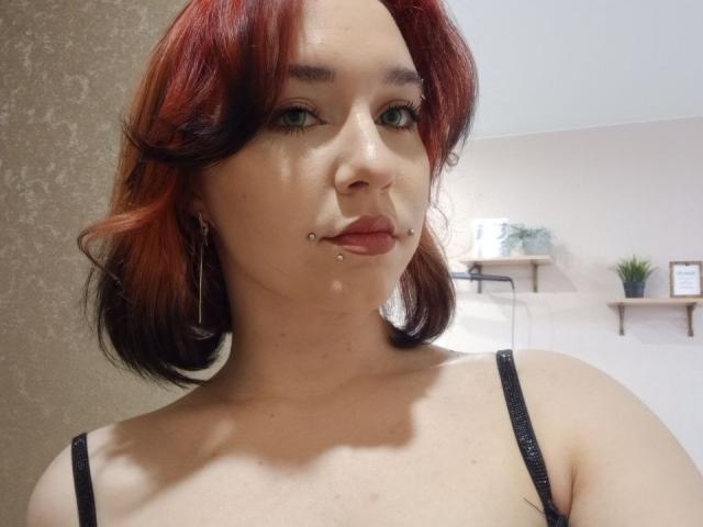 DinahLackey has red and black hair, green eyes, a lip piercing, and a black strap top, showing a clear face with slight water on her skin.