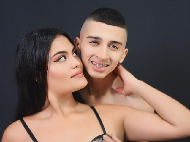 Mia, com cabelo preto, pele clara e maquiagem discreta, olha para a esquerda, enquanto Drony, com a cabeça raspada, pele clara e um piercing no nariz, sorri, tocando o pescoço dela..
