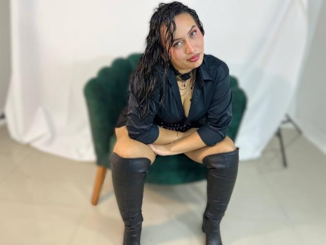 Amy Ross mit nassen, schwarzen Haaren, die ein schwarzes Hemd, eine Halskette und schwarze Stiefel bis zu den Knien trägt, sitzt auf einem grünen Samtstuhl und blickt mit einem leichten Lächeln nach oben..