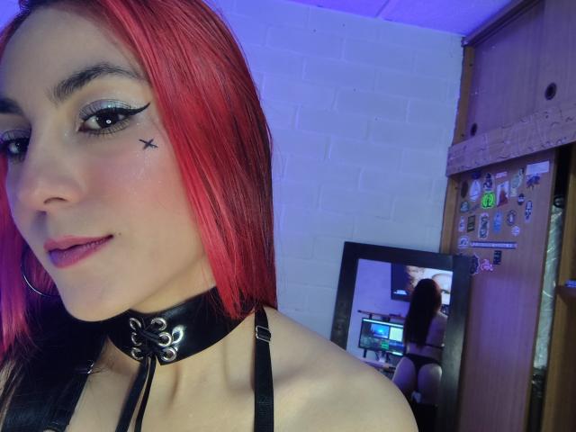 AmyStreetwise69 with red hair, black eyeliner, small star tattoo under eye, purple lipstick, black choker, black bra, mirror reflection showing her ass, in a room with blue lighting.