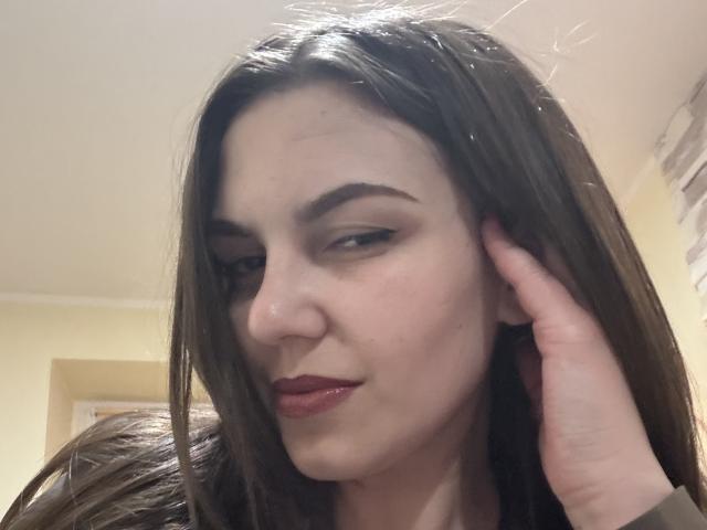 Kalipso69 has fair skin, dark brown straight hair, black eyeliner, red lipstick, and a hand touching her cheek. She looks down with a slight smile.