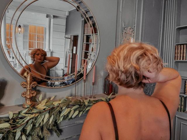 LorraSchmidt with short, wavy blonde hair, black strap top, looking in a large, round mirror, showing her back and reflection, arms raised, green leaf garland on gray shelf.