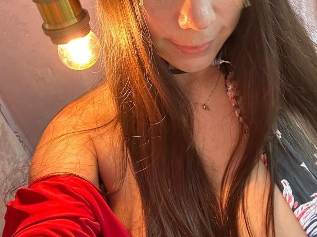FreyaraVenwo with long brown hair, slight smile, red velvet off-shoulder top, exposed shoulder, gold pendant, warm light from bulb, soft shadows, casual selfie, visible necklace, subtle makeup.