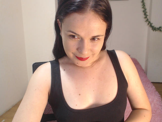 WendySchulz with pale skin, dark brown hair, red lipstick, black tank top, small tits, nose ring, sitting, slight smile, pink chair, wooden floor, leafy wall decoration.