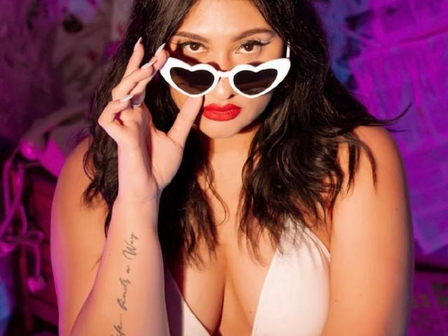 ElyRoss with heart-shaped white sunglasses, red lipstick, wavy black hair, white cleavage dress, tattoo on left forearm, purple and pink lighting. Boobs visible, hand near face.
