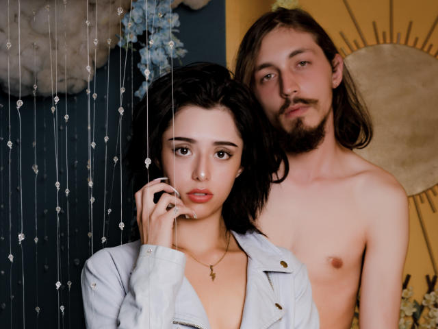JunnaAndKurt with dark hair and a nose ring, wearing a white jacket, has a slightly open shirt showing her tits. Kurt, shirtless, with long hair and a beard, stands behind.