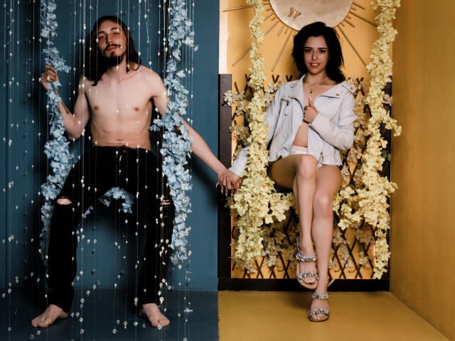 Shirtless JunnaAndKurt with black pants and water droplets, holding hands with white-jacketed JunnaAndKurt, legs crossed, sitting in floral background. JunnaAndKurt has tits, black hair, and a beard.