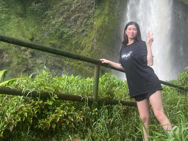 AnyuWu with long black hair, wearing a black T-shirt and black shorts, standing by a bamboo fence, making a peace sign, with a waterfall in the background.