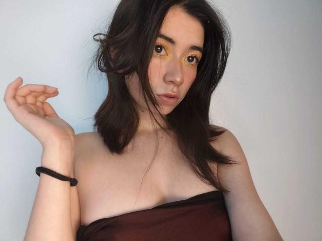 AnyuWu mit braunen Augen, gelbem Eyeliner und dunkelbraunem Haar, trägt ein trägerloses, braunes Oberteil und hebt ihre linke Hand. Am Handgelenk trägt sie ein schwarzes Armband..