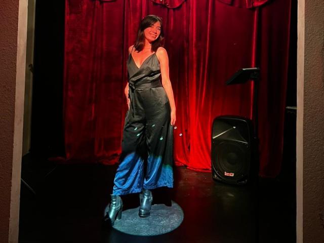 AnyuWu está de pie, vestida con un brillante vestido gris con degradado azul, y lleva botas azules metálicas, sonriendo. A su derecha hay un altavoz negro, y el fondo es una cortina roja, con un foco iluminándola..