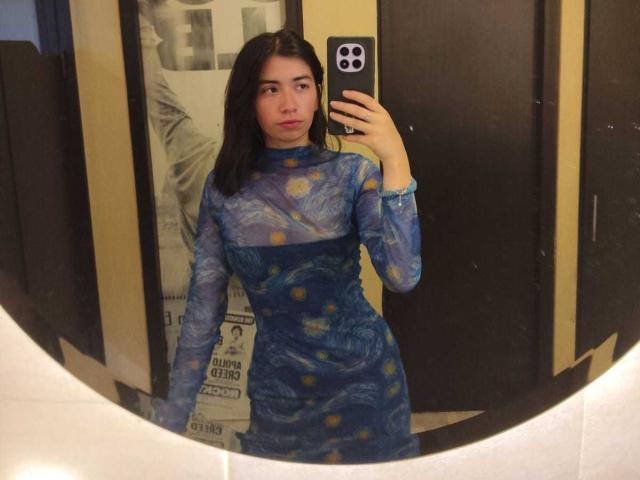 AnyuWu with dark hair, wearing a blue, long-sleeve, patterned dress, standing in front of a mirror, holding a phone, showing her reflection, with a neutral expression.