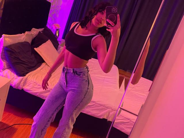 ChloeVixen, com cabelos pretos ondulados, vestindo uma regata preta e jeans de cintura alta, exibindo seus músculos abdominais e seios em uma selfie no espelho, segurando um telefone com iluminação roxa..