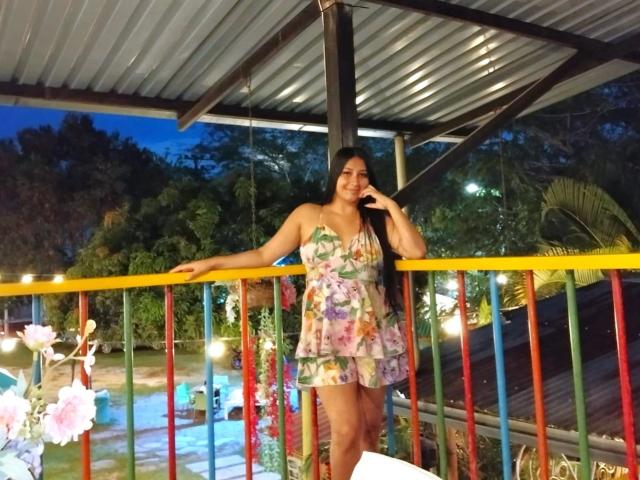 Catalina Rivero, con senos de tamaño medio, vestida con un vestido floral con volantes, está de pie en un balcón colorido, tocándose la mejilla, sonriendo a la cámara, bajo un techo de metal por la noche..