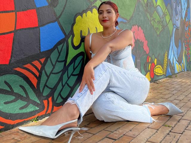 MiaBianchi with red hair, wearing a white strapless corset, light blue jeans, and white heels, sitting on a brick floor, showing her tits and ass in a graffiti wall.