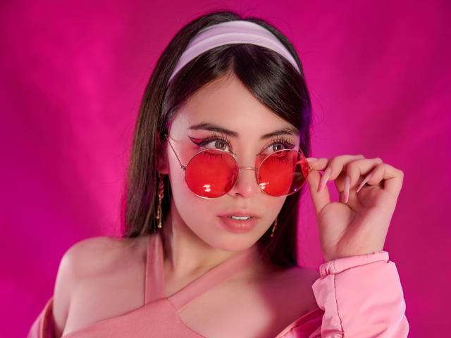 NoahAndRose with pink sunglasses, pink hairband, and pink off-shoulder top, holding her glasses, showing off her tits and pink nails, against a hot pink background.