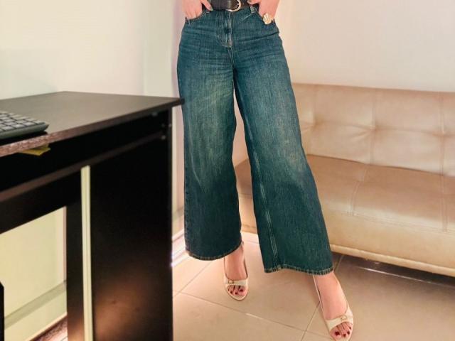 AdharaCooperr's blue denim pants, slightly flared, with a black belt, standing in heels, showing red-painted toenails, next to a dark desk and beige sofa.