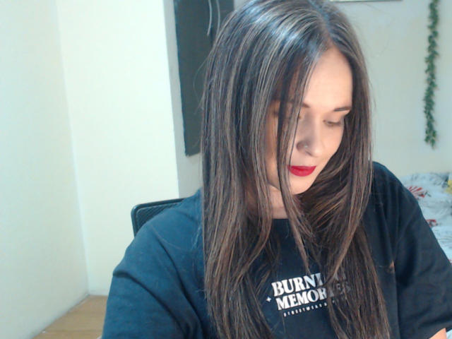 WendySchulz with long brown hair, red lipstick, and black shirt, looking down, her shiny hair slightly covering her face, in a bright indoor setting.