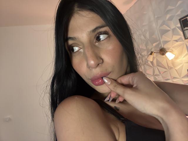 SophieBeaumont with black hair, brown eyes, nose ring, and pink lips, biting her finger, looking to the side, wearing a black top, visible shoulder tattoo.