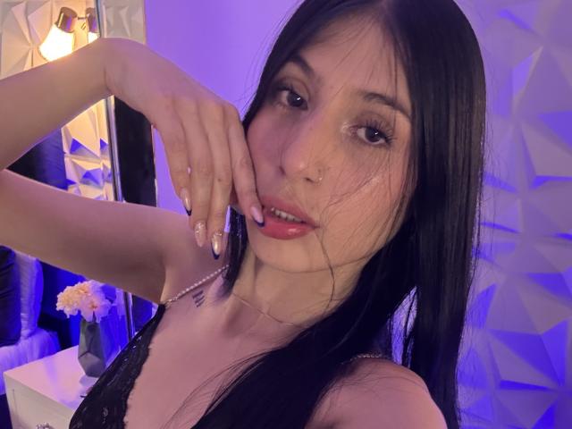 SophieBeaumont has long black hair, brown eyes, and a nose ring. She's touching her cheek with manicured nails, wearing black lace, and has pink lips.