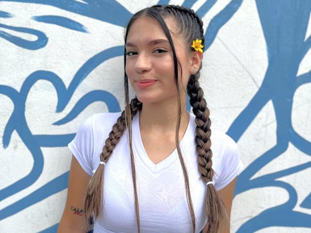 Milly Palmer ha lunghe trecce castane, un fiore giallo tra i capelli, una maglietta bianca a V e un tatuaggio sul braccio sinistro. Il suo seno piccolo è visibile. Sta sorridendo..
