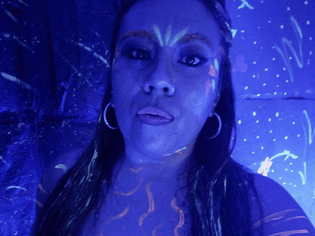 LexyDom with blue-tinted skin, glowing face paint, and hoop earrings, sticking out her tongue, has wet brown hair, and is wearing a low-cut top, showing her tits.