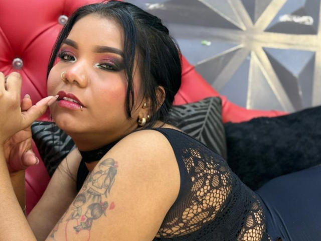 ElizabethSuaan with black hair, pink eyeshadow, nose ring, and tattoo on her arm, touching her pink lips with her finger. She's wearing a black lace top and gold earrings.