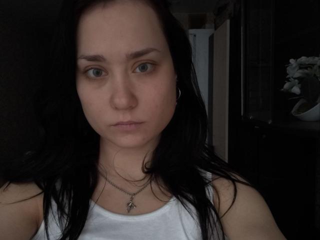 PatienceObremski with pale skin, black hair, green eyes, white tank top, small tits, silver necklace, serious expression, dim lighting, slightly messy hair, looking straight at camera.