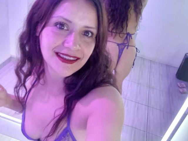 AriannaSeete with wavy brown hair, red lipstick, and purple bikini, showing her tits and ass in a mirror selfie, smiling with pinkish-blue lighting.