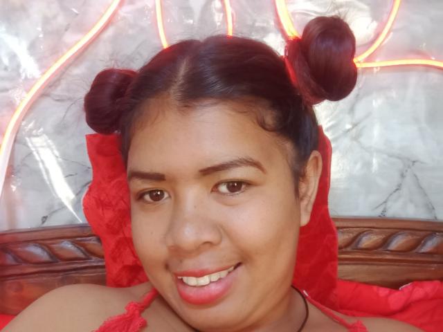 MariahSquirting69, avec des cheveux bruns foncés en deux chignons, un foulard rouge en dentelle, souriant avec du rouge à lèvres rose, des yeux marrons et une peau marron moyen, avec un appui-tête en bois en arrière-plan..