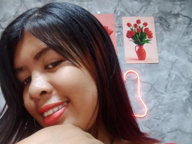 MariahSquirting69, souriant avec du rouge à lèvres, des cheveux noirs et lisses, et une peau brune, montrant son épaule, avec une lumière néon en forme de papillon rouge et un vase à fleurs rouges en arrière-plan..
