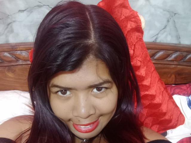 MariahSquirting69 tiene el cabello castaño oscuro, lápiz labial rojo y está sonriendo a la cámara, con un cojín rojo detrás de ella, acostada en una cama con cabecera de madera..