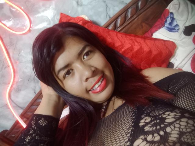MariahSquirting69, avec des cheveux violet foncé, souriante, portant un haut en dentelle noire, montrant de petites seins, allongée sur un lit avec un oreiller rouge, lumière néon en arrière-plan..