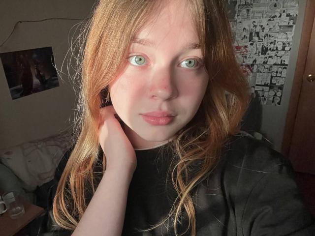 OzieBogg has green eyes, fair skin, and long reddish-brown hair. She's wearing a black shirt, with her hand touching her neck, looking into the camera.