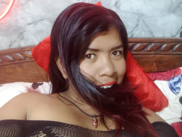 MariahSquirting69, con capelli scuri, occhi marroni e rossetto rosso, indossa una maglia nera in rete, guarda la fotocamera con un leggero sorriso, sdraiata su un letto con cuscini rossi..