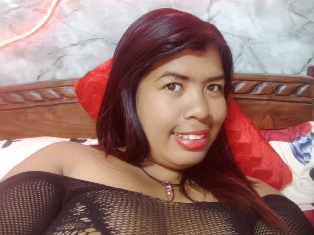 MariahSquirting69 with dark red hair, brown eyes, and red lipstick, wearing a black mesh top, smiling, showing her tits, red pillow behind her, wooden headboard, necklace with red gem.