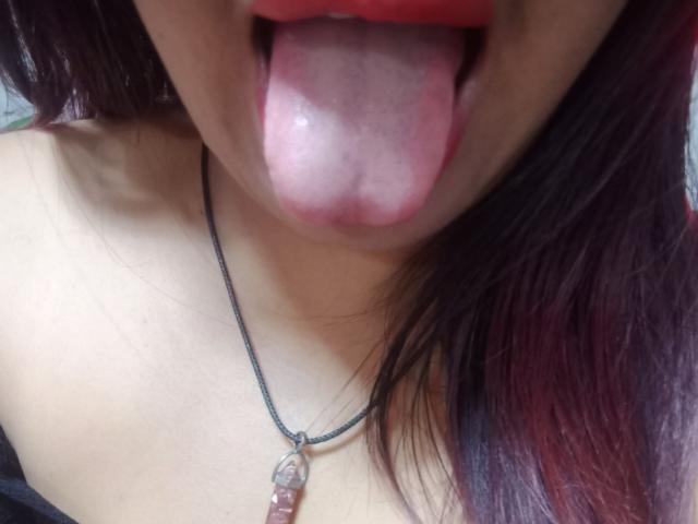 MariahSquirting69's tongue is out, sticky with white saliva, and she's wearing a black necklace with a pink stone. Her red lips are slightly parted.