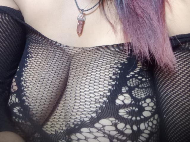 MariahSquirting69's tits through a black, fishnet, lace-patterned top, showing her nipples. She's wearing a necklace with a red, crystal pendant, and has pinkish, purple hair.