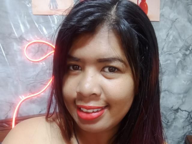 MariahSquirting69 with dark straight hair, medium brown skin, and red lipstick, smiling at the camera, neon red light in the background, showing her shiny nose and slightly flushed cheeks.