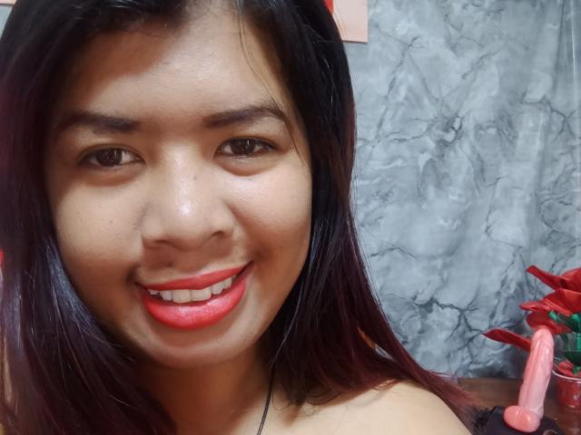 MariahSquirting69 tem olhos castanho-escuros, batom vermelho e cabelo preto na altura dos ombros, sorrindo para a câmera com um brinquedo sexual visível no lado direito da imagem..