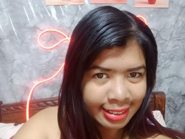 MariahSquirting69 tiene el cabello oscuro y brillante, ojos marrones y lápiz labial rojo. Está sonriendo y se pueden ver sus hombros desnudos. Una luz de neón roja está detrás de su cabeza..