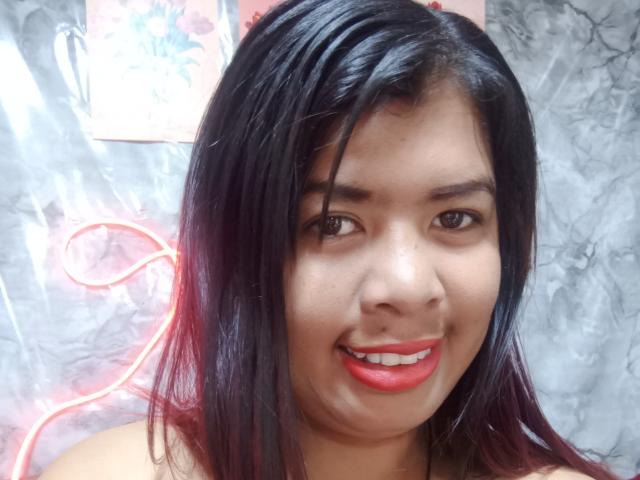 MariahSquirting69 a les cheveux noirs, les yeux marrons, et elle sourit avec du rouge à lèvres, montrant ses dents blanches. Elle porte un collier sur un fond à motifs de marbre..