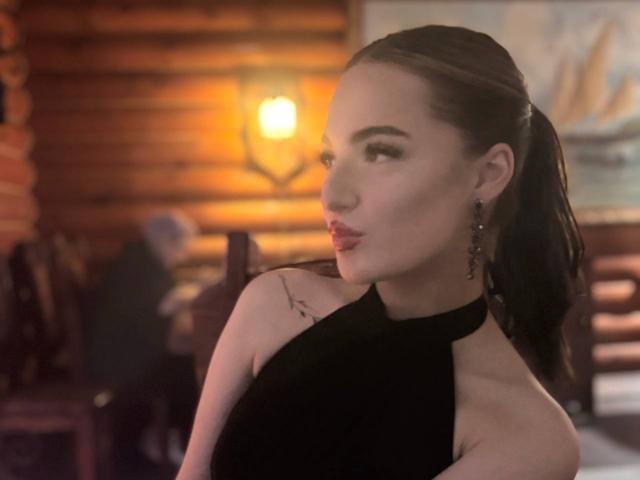MarietteHot with black hair in a ponytail, wearing a sleeveless black top, showing a tattoo on her right shoulder, looking over her shoulder with pouty lips.