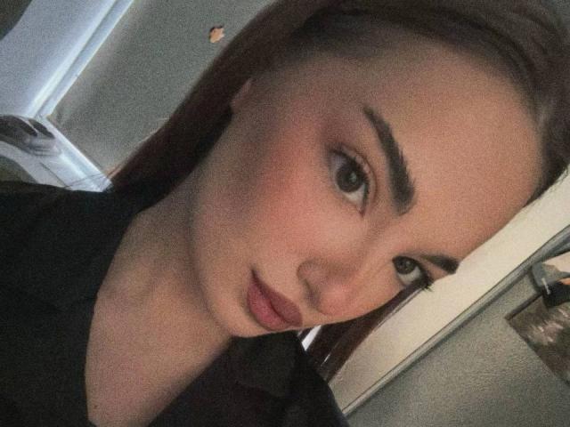 MarietteHot has brown eyes, thick eyebrows, and red lips. She's wearing a black shirt, with her brown hair pulled back. Her ass is not visible in the image.