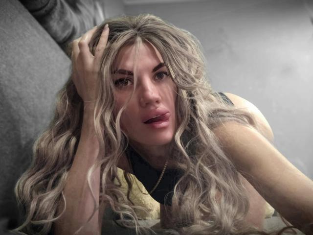 MilaLoona with long, wavy blonde hair, hand in her hair, looking up with parted pink lips, wearing a black top, gray couch in the background, soft light.