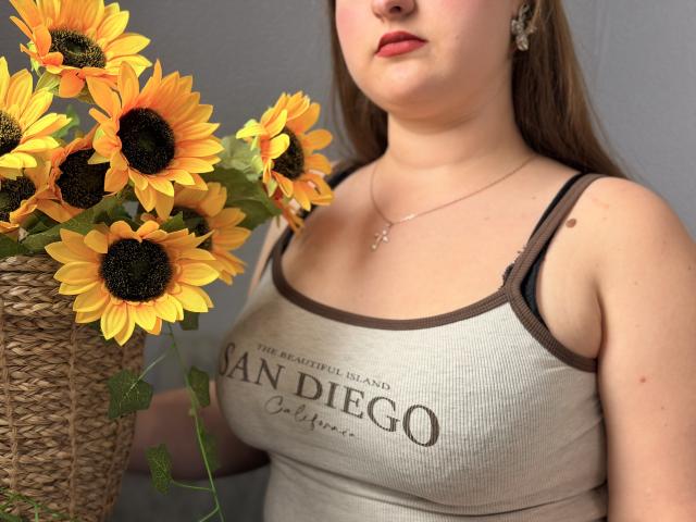 AvaNiss with big tits, wearing a grey tank top, holding a wicker vase with bright yellow sunflowers, standing near a grey wall, showing off her red lipstick.