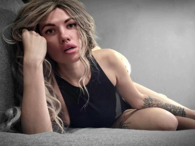 MilaLoona, con cabello rubio largo y ondulado, vestida con una camiseta negra, sentada en un sofá gris, luciendo cansada y con una mano en la cara, mostrando un tatuaje en su brazo..
