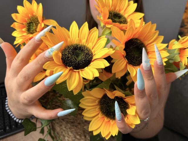 AvaNiss holding a bouquet of bright yellow sunflowers with her long, light blue, glittery, pointed nails. Her hands are in the foreground, showcasing her sharp, elegant nails.