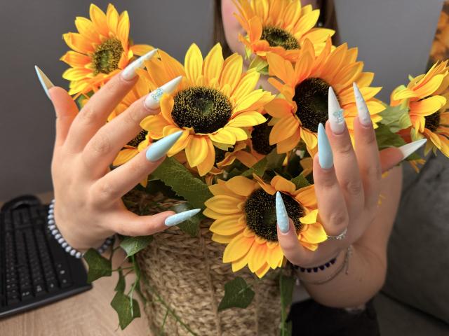 AvaNiss's hands with long, blue, glittery nails holding a bouquet of bright yellow sunflowers with dark centers, green leaves, and a woven basket.