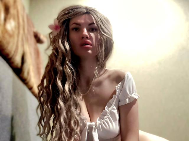 MilaLoona, avec ses cheveux blonds ondulés, une fleur rose, un haut blanc à épaules dénudées et un collier, regarde vers le haut avec les lèvres légèrement entrouvertes, mettant en valeur sa petite poitrine. Lumière douce, légèrement floue..