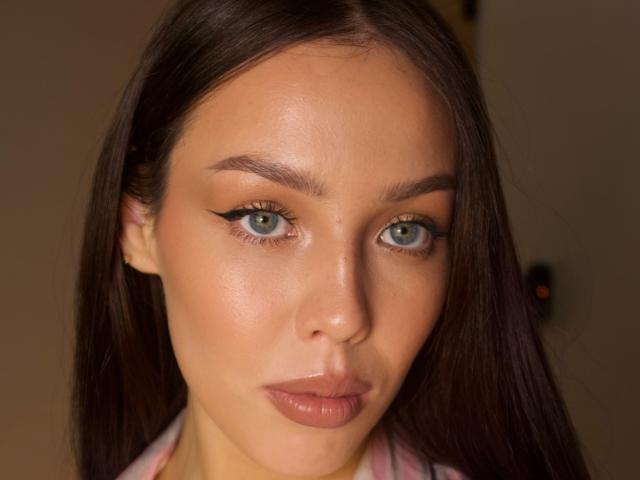 JaneWow has shiny green eyes, thick black eyeliner, full pink lips, and smooth tan skin. Her brown hair is straight and she's staring directly at the camera.